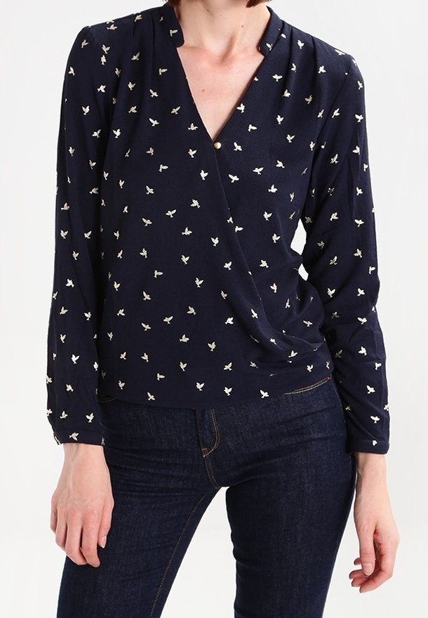 Femme portant un chemisier à manches longues bleu marine avec un petit motif d'oiseaux blancs et un jean ajusté bleu foncé, debout les mains détendues.