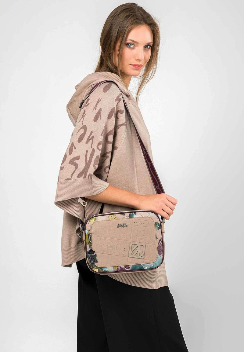 Bolso bandolera beige con acentos florales y diseño texturizado. Presenta un cierre de cremallera y correa de hombro ajustable. Forma rectangular estructurada.