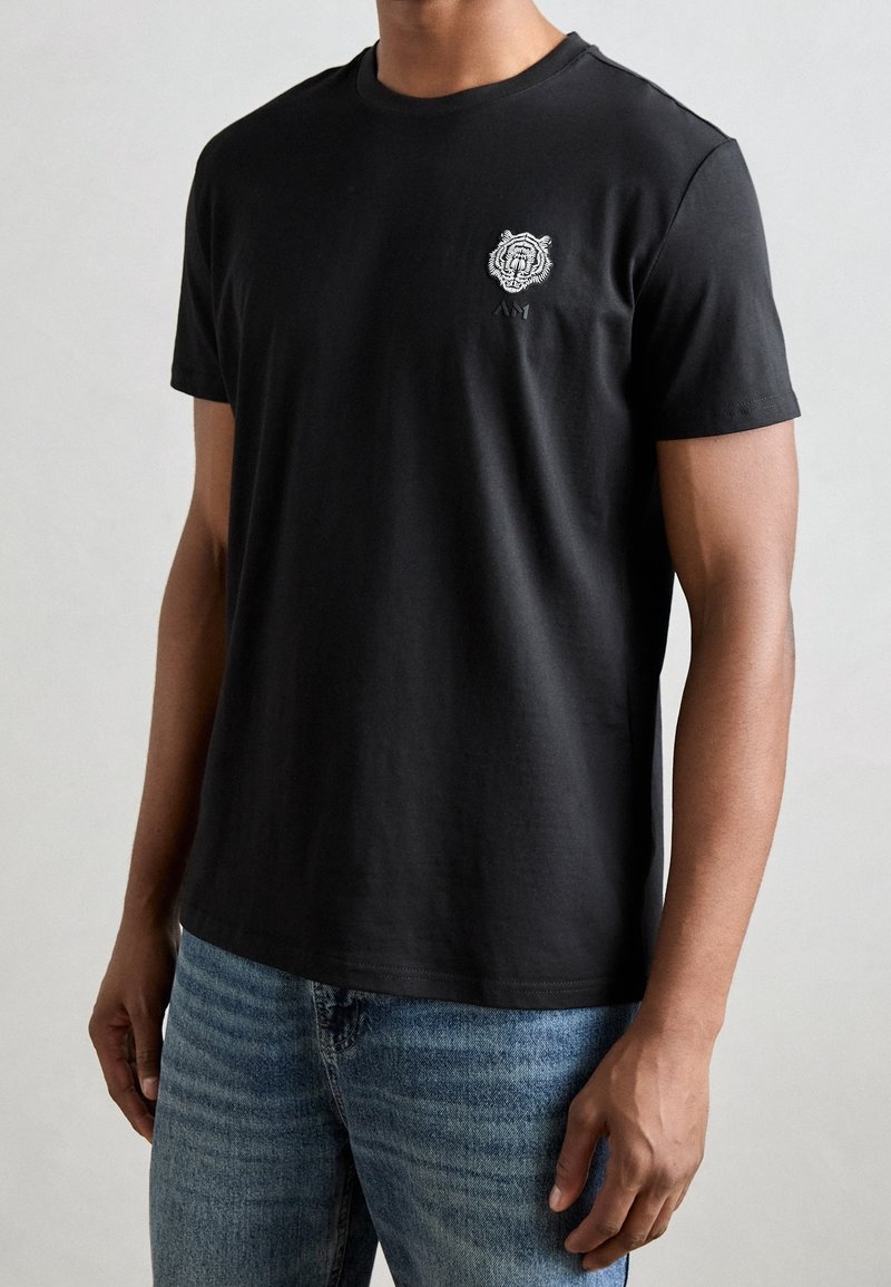Black cotton t-shirt with short sleeves, crew neck, and a small white lion graphic on the left chest. Paired with light blue jeans.