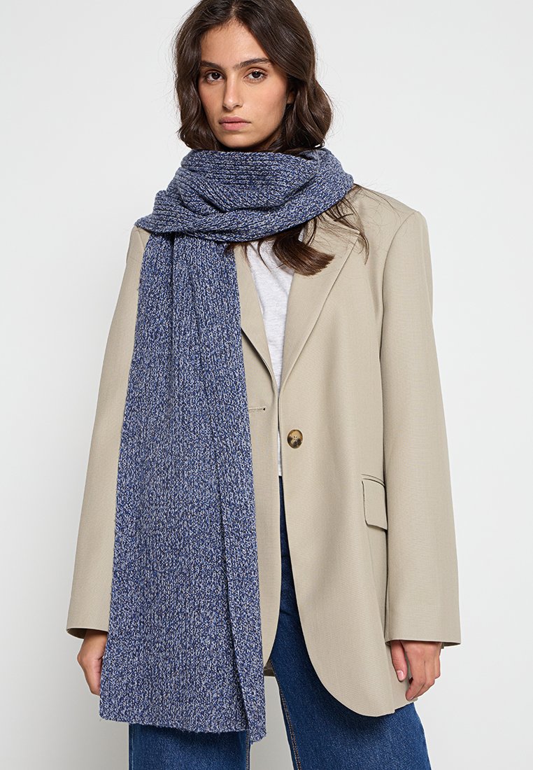 Écharpe en tricot bleu avec une finition texturée drapée autour du cou. Portée sur un blazer beige clair, complétant l'ensemble décontracté.