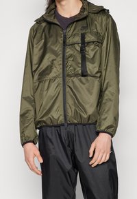 Olivgrön lätt vindjacka med dragkedja framtill, justerbar huva och svarta dragkedjedetaljer; har bröstficka med rem.