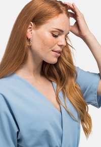 Orecchini a cerchio dorati con accenti circolari, abbinati a una blusa avvolgente azzurro chiaro con maniche corte. Tessuto morbido e vestibilità rilassata.