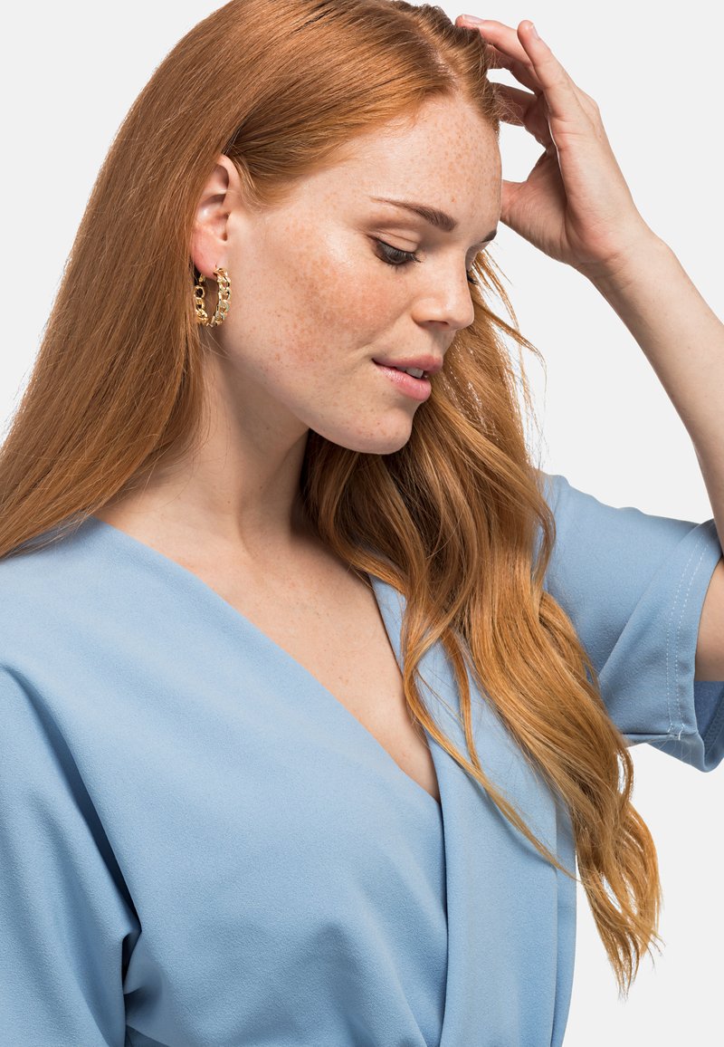 Orecchini a cerchio dorati con accenti circolari, abbinati a una blusa avvolgente azzurro chiaro con maniche corte. Tessuto morbido e vestibilità rilassata.