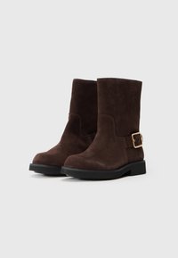 Botas de tobillo de ante marrón con una suela de goma negra y detalle de hebilla dorada en el costado. Diseño recto y estructurado con una textura suave.