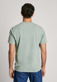 Šviesiai žalia medvilnėms marškinėliai trumpomis rankovėmis, apvalia iškirpte ir laisvu siluetu. Paskes gale matosi lygi tekstūra ir minimalūs dizaino akcentai.