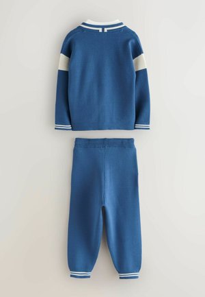 Blauwe gebreide trui met witte strikaccenten op de mouwen en kraag, gecombineerd met bijpassende blauwe broeken. Zachte textuur, casual ontwerp.