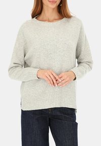 Kobieta w jasnoszarym dzianinowym swetrze z długimi rękawami i ściągaczem na dole, w ciemnoniebieskich dżinsach, z rękami złożonymi na wysokości talii.