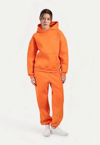 Pomarańczowa bluza z kapturem i dopasowane spodnie dresowe. Miękki materiał, luźny krój, kieszeń kangurka, ściągacze na rękawach i nogawkach. Połączone z białymi butami sportowymi.