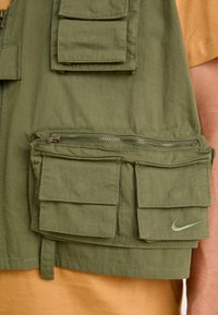 Ελαιόχρωμο γιλέκο με πολλαπλές μεγάλες μπροστινές τσέπες, μία τσέπη με φερμουάρ και το λογότυπο της Nike. Κατασκευασμένο από αναπνεύσιμο υλικό, διαθέτει καθαρό, χρηστικό σχεδιασμό.