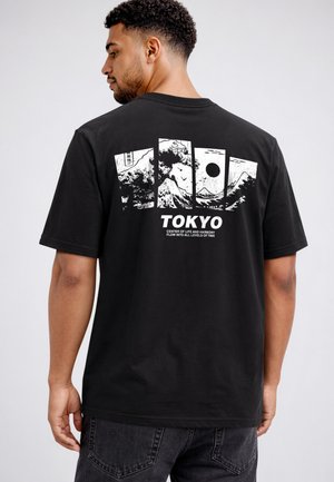 Man wearing black T-shirt with white design of Mount Fuji, waves, and sun, labeled "TOKYO" with text about life and harmony.