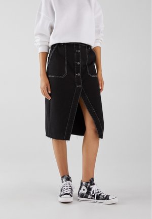 Person, der bærer et sort midi-nederdel med knapper foran med hvide syninger, en hvid sweatshirt og sort-hvide ternede høje sneakers.