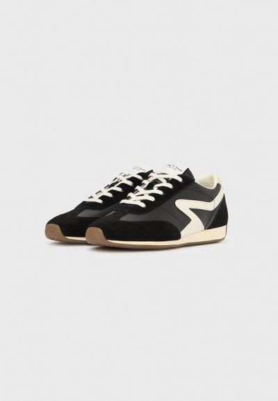 Fekete és krémszínű sportcipők, melyek művelúr és bőr dizájnnal, klasszikus formával, kerek orral és kontrasztos cikk-cakk mintával rendelkeznek az oldalukon.