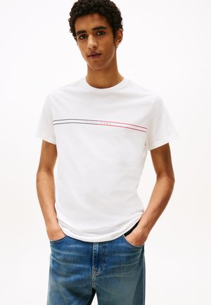 T-shirt bianco in cotone con maniche corte, caratterizzato da una banda grafica orizzontale in blu e rosso, con il testo "TOMMY JEANS" al centro.