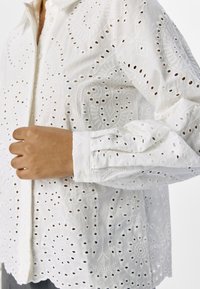 Blouse blanche brodée avec des motifs découpés, un front à boutons, et des manches longues, fabriquée en tissu léger avec un ourlet festonné.