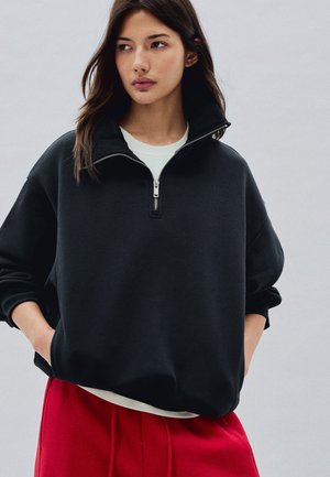 Jonge vrouw met lang donker haar draagt een zwart halflangs rits sweatshirt over een wit overhemd en rode broek, handen in de zakken, kijkt opzij.