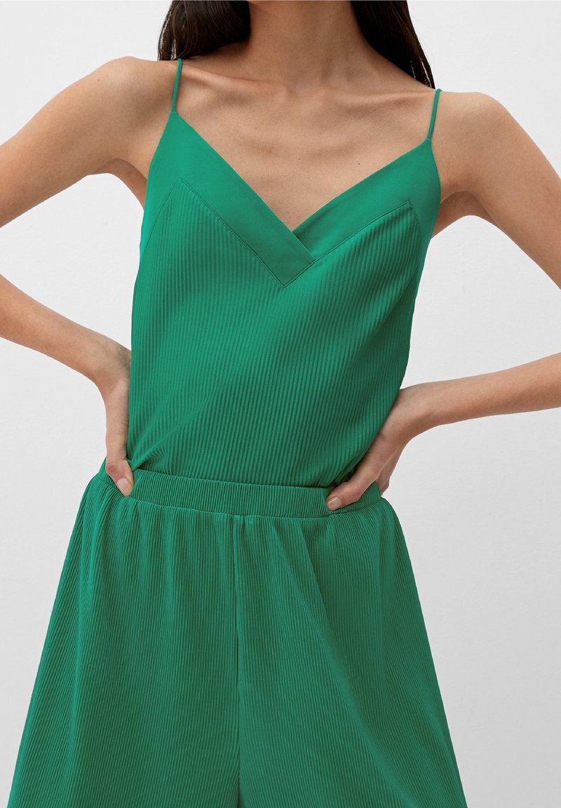 Frau trägt ärmelloses grünes, geripptes Top mit V-Ausschnitt und passende, hochtaillierte Hose, Hände auf die Hüften gestützt, einfarbiger heller Hintergrund.