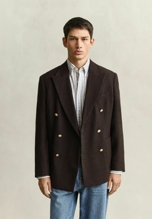Junger Mann steht, trägt ein dunkles, zweireihiges Sakko mit goldenen Knöpfen, ein gestreiftes Hemd und blaue Jeans vor einem schlichten Hintergrund.