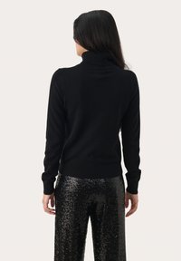 Melna turtlenecks džemperis ar piegulīgu piegriezumu un ribotām piedurknēm, apvienots ar melnām spīguļojošām biksēm. Gluda tekstūra un minimāli rotājumi.