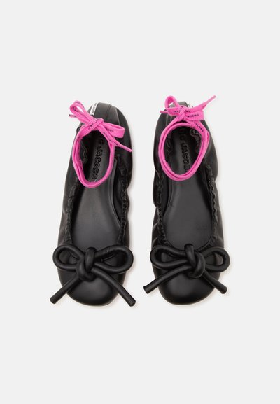 Paire de ballerines noires avec de grands nœuds noués sur les orteils et des rubans roses vifs noués autour des chevilles sur fond blanc.