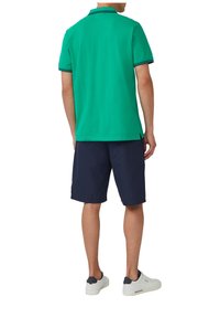 Grünes Poloshirt mit kurzen Ärmeln und einem Kragen, kombiniert mit dunkelblauen Shorts. Das Shirt hat einen kontrastierenden Besatz am Kragen und an den Ärmeln.