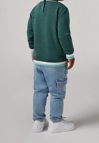 Grüner Sweatshirt mit Rundhalsausschnitt und weißen sowie türkisfarben gestreiften Bündchen, kombiniert mit hellblauen Cargo-Jeans und weißen Sneakern. Rückansicht.