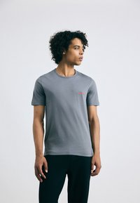 T-shirt gris à manches courtes avec col rond, comportant un petit logo rouge sur la poitrine gauche. Matière en coton doux, design ajusté.