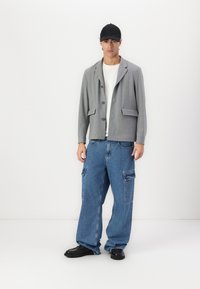 Siv blazer s dvema sprednjima žepoma, oblečen preko bele majice. Ohlapne modre denim hlače z žepi s strani. Črna kapa in čevlji.