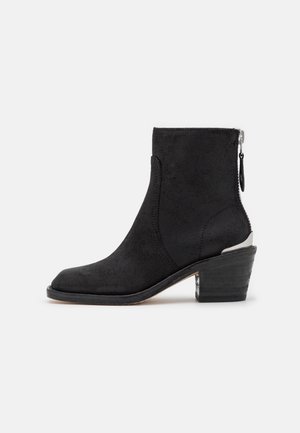 Bottine en daim noir avec talon bloc, fermeture éclair latérale et détail métallique sur le talon, sur fond blanc.