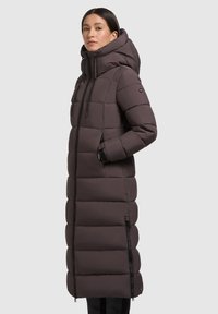 Dolga, puhasta bunda temno rjave barve z visokim ovratnikom in veliko kapuco. Ima zadrge, žepe ter značilne navpične prešite odseke.