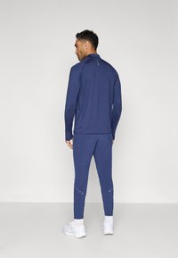 Marineblaue sportliche Jacke und passende Hose aus glattem Stoff, mit langen Ärmeln, hohem Kragen und reflektierenden Akzenten. Weiße Turnschuhe vervollständigen den Look.