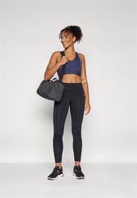 Zwarte leggings, marineblauwe sportbeha en zwarte sneakers. Draagt een donkergrijze sporttas. Glad, strak weefsel met duidelijke naden.
