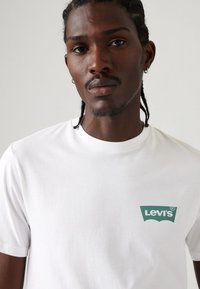 Mann med flettede hår og øreringer iført en hvit Levi's t-skjorte med grønn logo på brystet mot en ensfarget bakgrunn.