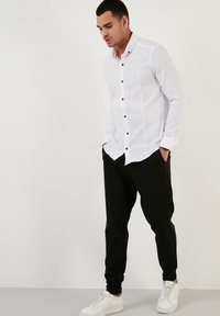 Hvid skjorte med lange ærmer og sorte knapper, parret med sorte joggerbukser og hvide sneakers. Glat stof.