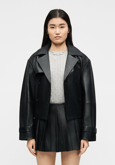 Junge Frau mit langen dunklen Haaren, die eine schwarze Lederjacke, einen grauen Zopfpullover, einen schwarzen plissierten Lederrrock und eine silberne Halskette trägt.