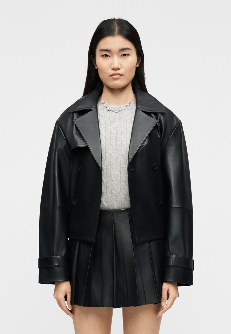 Νεαρή γυναίκα με μακριά σκούρα μαλλιά που φοράει μαύρο δερμάτινο μπουφάν, γκρι πουλόβερ με πλεκτό σχέδιο, μαύρη δερμάτινη πλισέ φούστα και ασημένιο κολιέ.