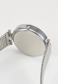 Metalinis laikrodis su apvaliu sidabriniu korpusu ir tinkleliu iš nerūdijančio plieno. Šone esantis mygtukas, blizgi paviršius ir minimalistinis dizainas.