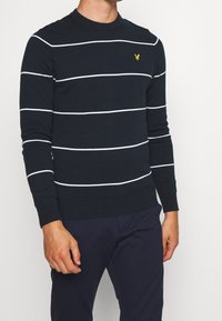 Pull rayé bleu marine en matière tricotée. Présente des rayures blanches horizontales et un petit emblème jaune sur la poitrine. Encolure ronde.