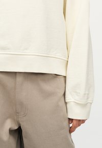 Κοντινό πλάνο ενός ατόμου που φοράει φούτερ σε κρεμ απόχρωση και μπεζ παντελόνι, εμφανίζοντας το κάτω μέρος του κορμού και το δεξί χέρι χαλαρό στο πλάι.