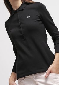 Polo à manches longues noir en tissu doux, avec une patte de boutonnage et un petit logo brodé blanc sur la poitrine.