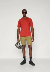 Červená košeľa s krátkym rukávom, khaki šortky, čierne ponožky a čierne topánky. Drží sivú prilbu. Jednoduchý dizajn s minimálnymi akcentmi.