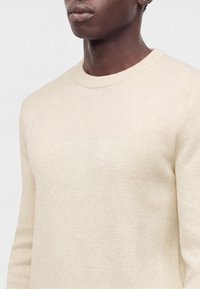Gros plan d'une personne portant un pull en maille côtelée crème à manches longues avec un col rond.