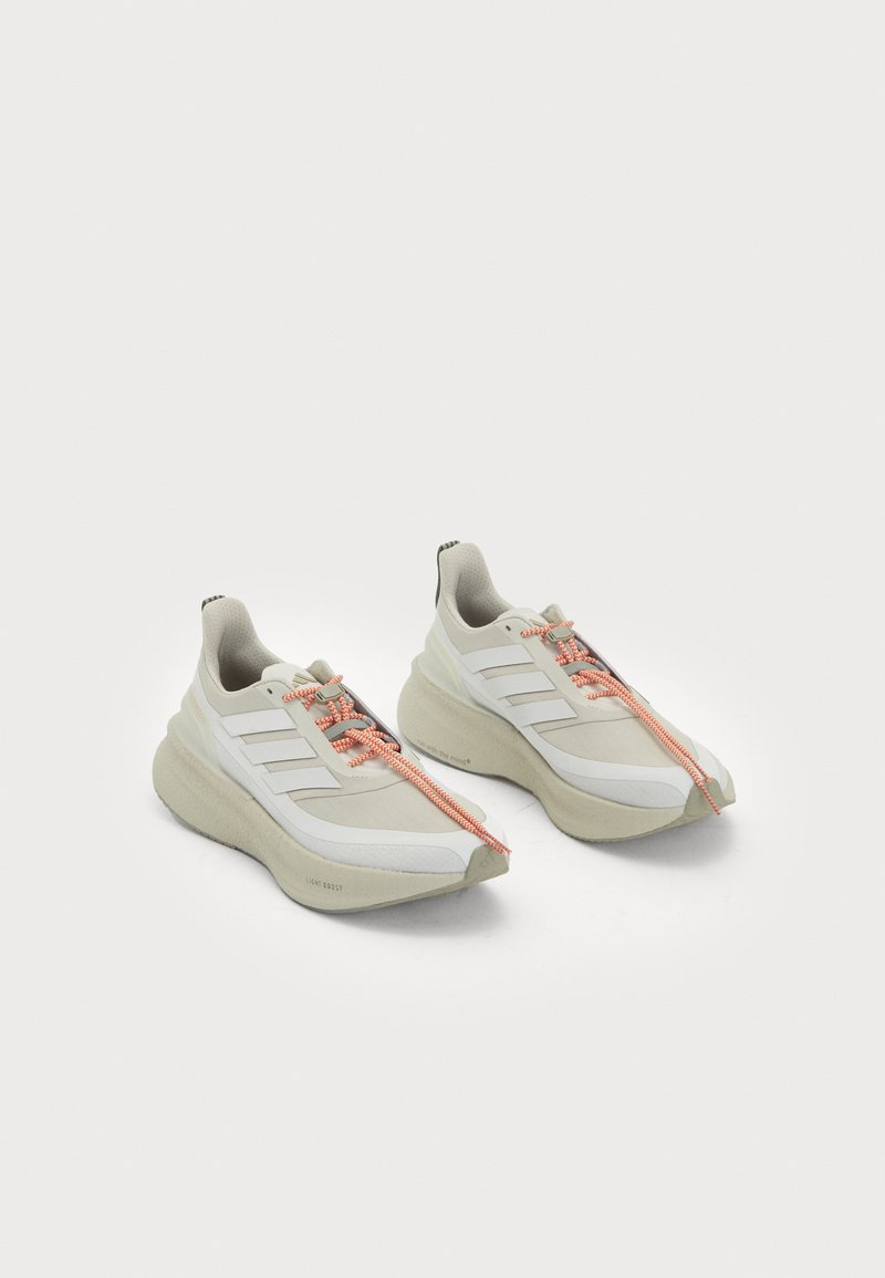 White athletic shoes with a textured upper, featuring three diagonal stripes, orange laces, and a cushioned, curved sole design.