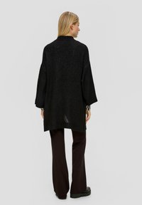 Schwarzer, oversized Strickpullover mit hohem Kragen, fallenden Schultern und lockerer Passform. Weiche Textur mit leichtem Glanz. Kombiniert mit dunklen, weiten Hosen.