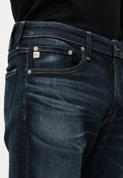 Σκούρο μπλε τζιν παντελόνι με ορατή μπροστινή τσέπη, κουμπιά ριβέτες, ραφές και μικρή λευκή ετικέτα με κόκκινο λογότυπο "AG".