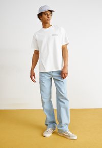 T-shirt en coton blanc avec logo brodé, jeans en denim bleu clair et baskets beiges. Le modèle porte une casquette à rayures claires.
