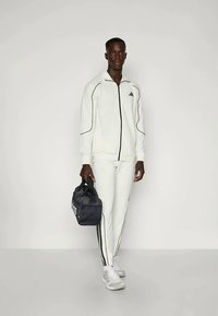 Biely adidas športový oblek s čiernymi akcentmi, obsahujúci zip-up bundu a zodpovedajúce nohavice. Model drží čiernu športovú tašku.
