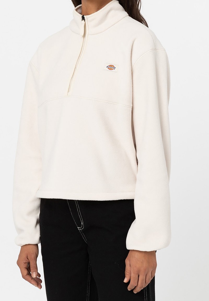 Pullover in pile color crema con colletto a mezza zip, dettaglio di cucitura frontale e una piccola etichetta con logo sul petto. Vestibilità comoda e maniche lunghe.