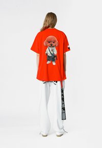 Camiseta roja de gran tamaño con un gráfico de un perro de dibujos animados en la parte trasera, combinada con pantalones blancos que tienen acentos de texto negro y un detalle de cinturón.