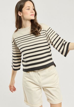Femme portant un haut à rayures horizontales beige et noir avec des manches aux coudes et un short beige retroussé, regardant vers sa gauche.