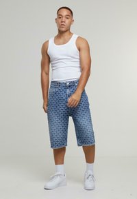 Pantalones cortos de mezclilla azules con un patrón geométrico, con un corte recto, dobladillos enrollados y cierre de botón, combinados con una camiseta sin mangas blanca y zapatillas.